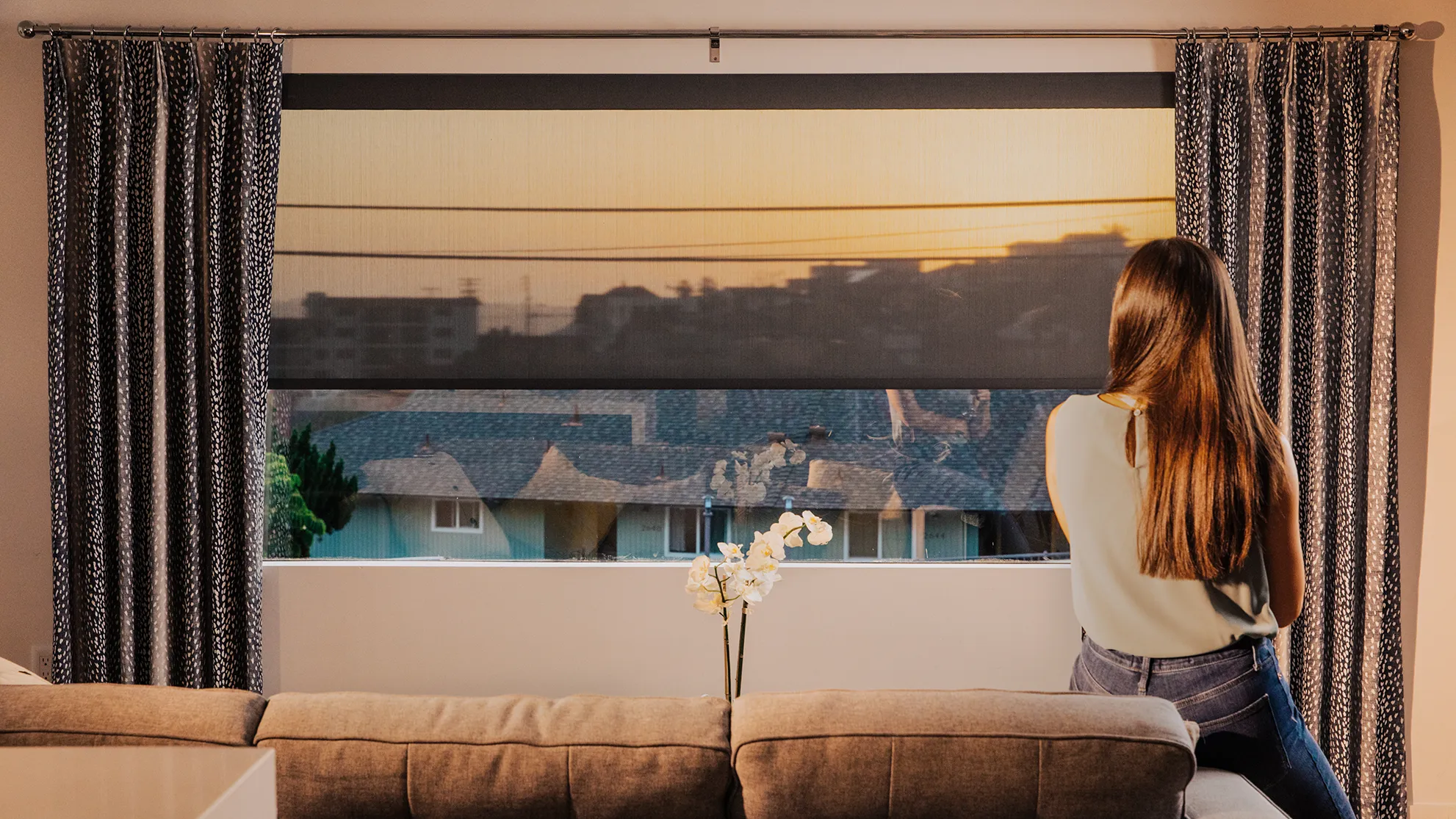 Woman sitting on a sofa in a living room with roller shades layered beneath custom pinch pleat curtains